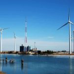 Trump AXES Massive Wind Project — Grid DISASTER Averted Wind turbines and a communication tower near a body of water under a clear blue sky