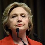 Hillary’s Shock Approval of Trump’s Middle East Move Woman in red jacket speaks into two microphones.