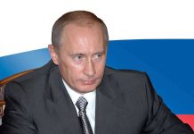A serious-looking man in a suit sitting at a table with a Russian flag in the background
