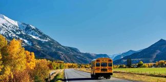 A yellow school bus driving on a rural road surrounded by autumn trees and mountains