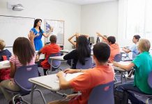 America’s Classrooms Hit By Major Shift Teacher in a blue dress instructing students in a classroom with hands raised