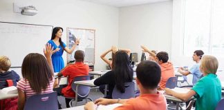 Teacher in a blue dress instructing students in a classroom with hands raised