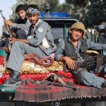 Three armed soldiers sitting on the back of a military vehicle in an urban setting