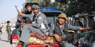 Three armed soldiers sitting on the back of a military vehicle in an urban setting