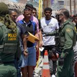 $5.7B In Migrant Aid SLASHED Border patrol agents inspecting group of individuals in line.