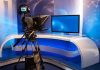 Journalists NOW in Danger Zones – Who Pays? TV studio with camera and empty anchor desk.