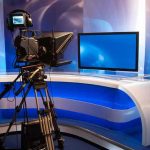 Journalists NOW in Danger Zones – Who Pays? TV studio with camera and empty anchor desk.