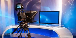 Journalists NOW in Danger Zones – Who Pays? TV studio with camera and empty anchor desk.