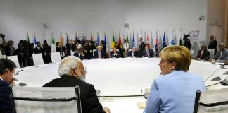Mystery Iran Deal Stuns G7 People seated around a large round table, flags behind.