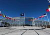 Europe’s Future? Americans Stuck With The Bill Flags outside NATO headquarters building under clear blue sky.
