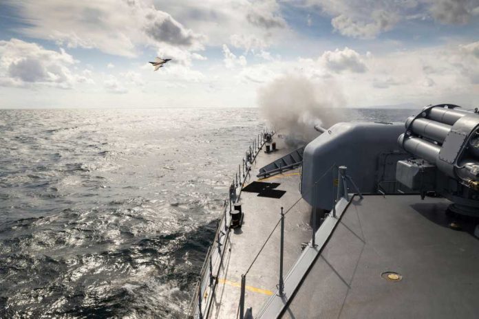 shutterstock_2129183807 Naval ship firing a cannon with a fighter jet flying overhead