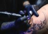 Tattoo Tax Panic Explodes Online Tattoo artist with black gloves inking a detailed arm design