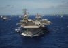 Hormuz Standoff Explodes—Oil On Edge An aircraft carrier surrounded by a fleet of naval ships in the ocean