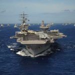 Hormuz Standoff Explodes—Oil On Edge An aircraft carrier surrounded by a fleet of naval ships in the ocean