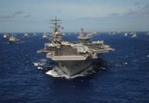 Hormuz Standoff Explodes—Oil On Edge An aircraft carrier surrounded by a fleet of naval ships in the ocean