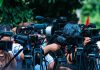 DHS Blames Biden For Preventable Killing TV news cameras crowding together at an outdoor event