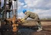 Cyberattack HITS Oil Tanker Headed for Texas Worker in blue hard hat operating rusty oil pump equipment outdoors