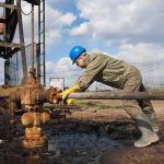 Cyberattack HITS Oil Tanker Headed for Texas Worker in blue hard hat operating rusty oil pump equipment outdoors