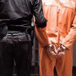 ‘Transgender’ Influencer Arrested on Child Molestation Allegations Prisoner in orange jumpsuit handcuffed with guard standing beside