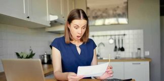 Woman in kitchen reacting to a bill at a table with laptop and calculator