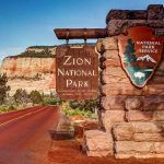 They Called It A Hiking Accident For 20 Years Zion National Park entrance sign beside red road and sandstone cliffs