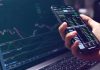 Why New York Is Coming After Polymarket Hand holding smartphone with stock market app beside laptop screen with charts