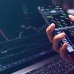 Why New York Is Coming After Polymarket Hand holding smartphone with stock market app beside laptop screen with charts