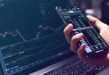 Why New York Is Coming After Polymarket Hand holding smartphone with stock market app beside laptop screen with charts