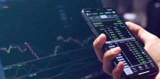 Why New York Is Coming After Polymarket Hand holding smartphone with stock market app beside laptop screen with charts