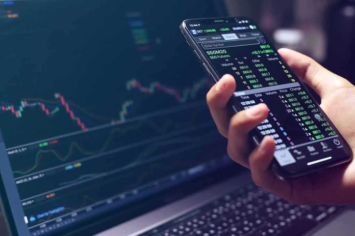 shutterstock_1728912097 Hand holding smartphone with stock market app beside laptop screen with charts