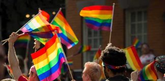 School Makes BOLD ‘LGBTQ+’ October Declaration Bear Pascoe at a pride parade holding a rainbow flag among a cheering crowd