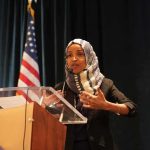Omar’s Allies Call Her Raid Political PAYBACK Ilhan Omar speaking at a podium with a microphone