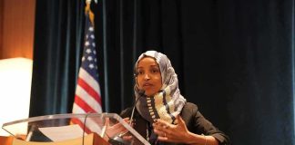 Omar’s Allies Call Her Raid Political PAYBACK Ilhan Omar speaking at a podium with a microphone