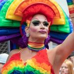 Drag performer in vibrant rainbow costume raising an arm at a parade