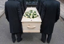 Four people carrying a coffin with white flowers