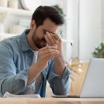Man rubbing eyes holding glasses near laptop