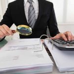 Person examining documents with magnifying glass and calculator.