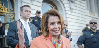 Woman in orange blazer speaking surrounded by security personnel