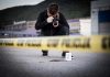 Crime scene photographer behind police tape with evidence marker.