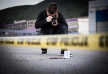 Crime scene photographer behind police tape with evidence marker.