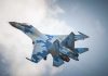 A blue camouflage fighter jet performing an aerial maneuver against a cloudy sky
