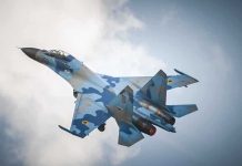 A blue camouflage fighter jet performing an aerial maneuver against a cloudy sky